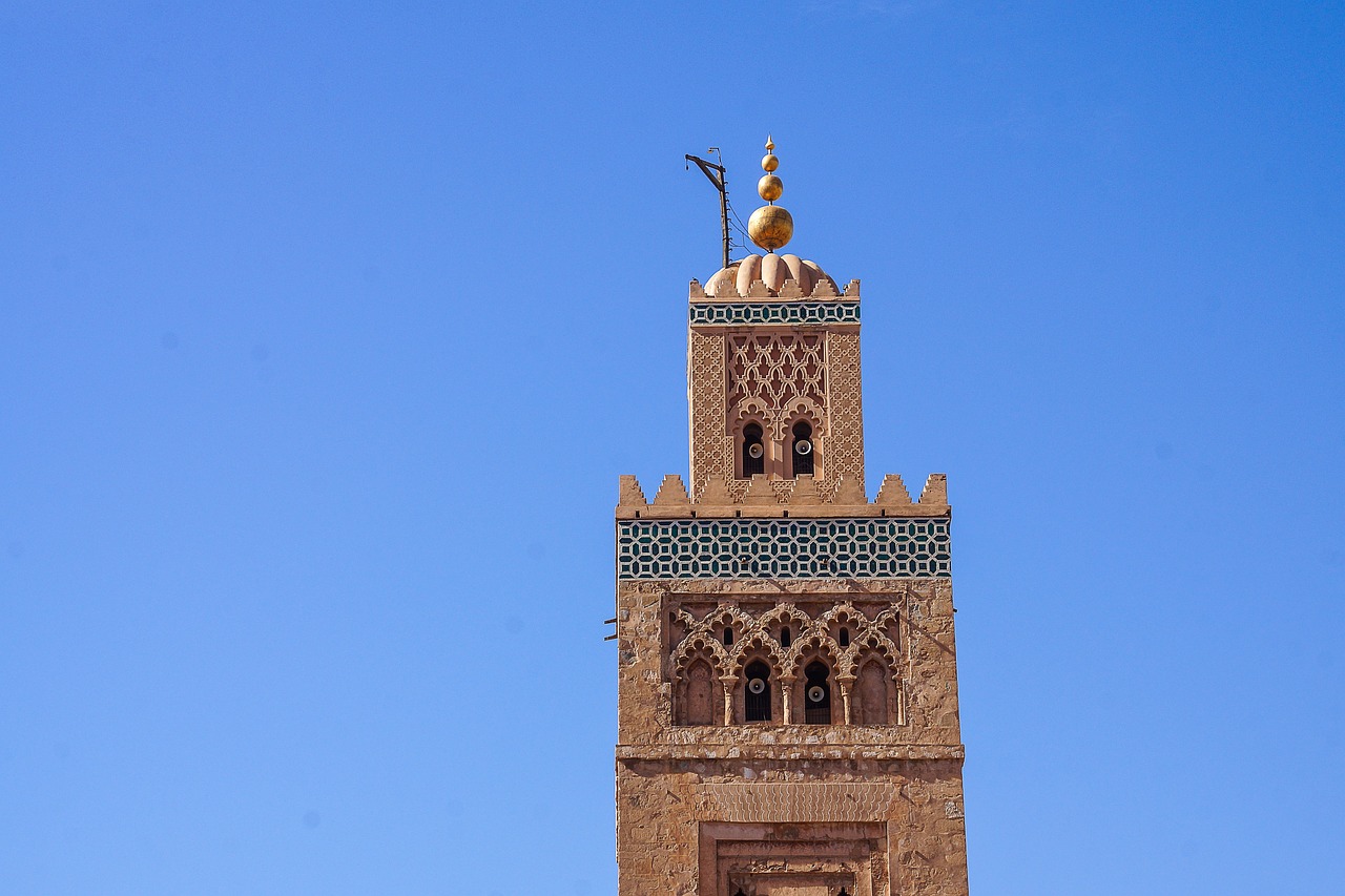 Questa fotografia ritrae la parte superiore di un alto minareto decorativo contro un cielo azzurro terso. Il minareto è composto da pietra bruno-rossastra, con elaborate piastrelle verdi e blu sulle parti più alte e un pinnacolo dorato in cima. Questo stile è comune nell'architettura islamica marocchina e si tratta di una moschea a Marrakech.