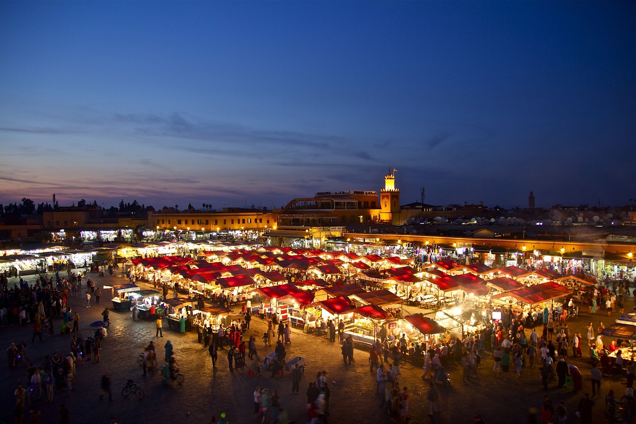 Questa è una scena notturna dinamica e animata, ripresa da una vasta piazza pubblica, molto probabilmente Djemaa el-Fna a Marrakech. La piazza è illuminata da innumerevoli luci provenienti da bancarelle di cibo e attività di mercato, che creano un incantevole chiarore sulla vasta folla. Il cielo sopra di noi è di un blu intenso, simile a quello del crepuscolo, con alcune strutture visibili in lontananza.